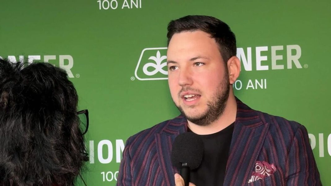 Man in a striped blazer speaks into a handheld microphone during an interview against a green branded backdrop (shows '100 ANI').