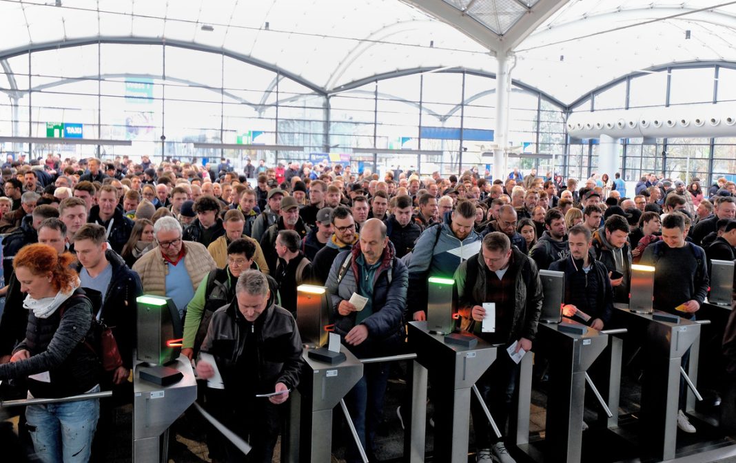 Large crowd of travelers waits at security gates with green and orange indicator lights in a glass-walled transit hub or station.