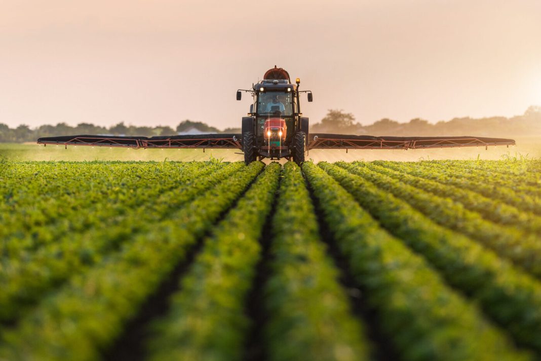 Tractor with a wide spraying boom in a green field at sunrise, applying liquid to crops