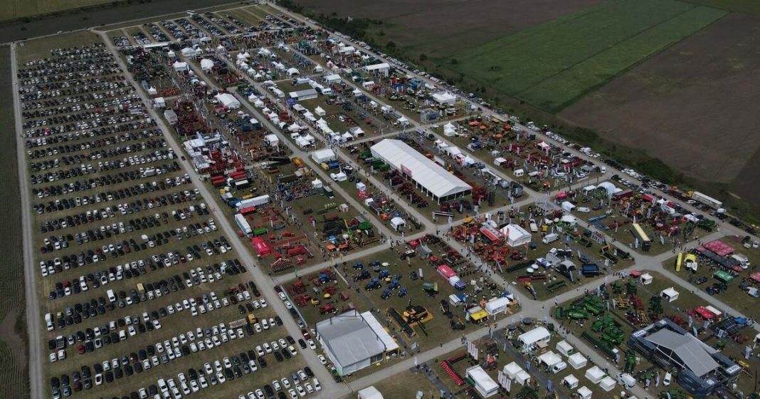 Aerial view of a large outdoor exhibition with rows of cars, tents, and machinery on display, spanning a wide fairground.