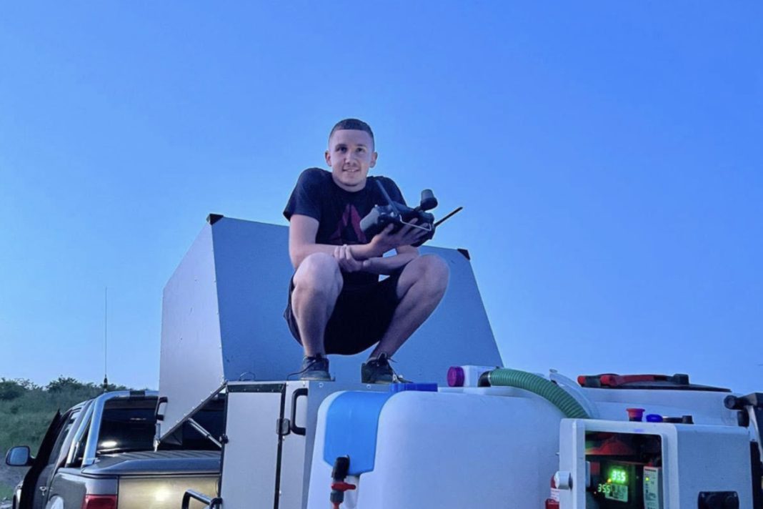 Man crouching on a metal platform atop a service truck, holding a drone controller outdoors.
