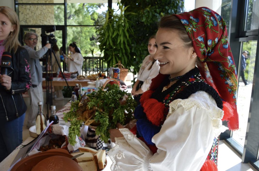 Smiling woman in traditional folk dress with a red floral headscarf offers fresh herbs at a table of bread and food during a cultural event; journalists film in the background.