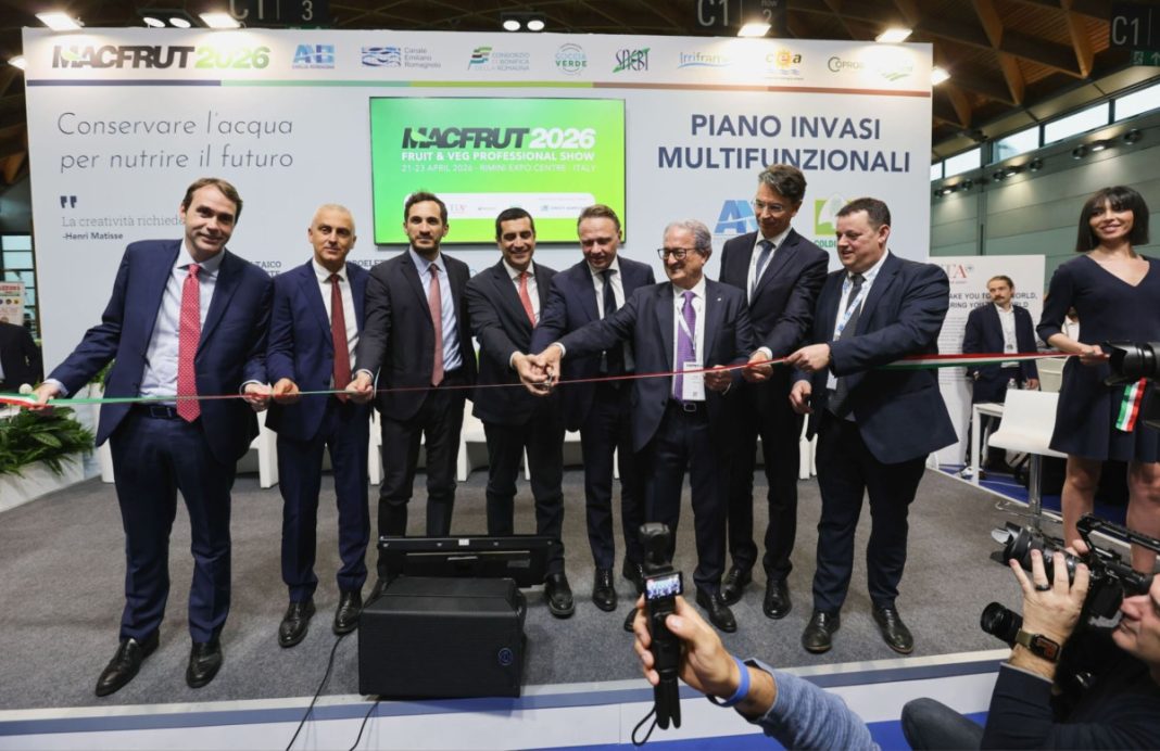 Eight men in suits cut a red ribbon at a NACFRUT 2026 booth as photographers film, with Italian banners in the background.