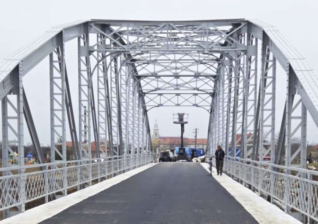Steel truss bridge with a paved pedestrian path, person standing with a bicycle on the right, city buildings in the background.