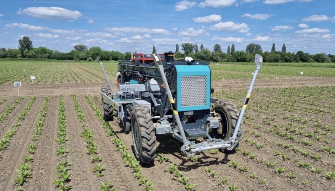 Autonomous farming robot with blue housing and lifting arms in a sunlit field of young crops.
