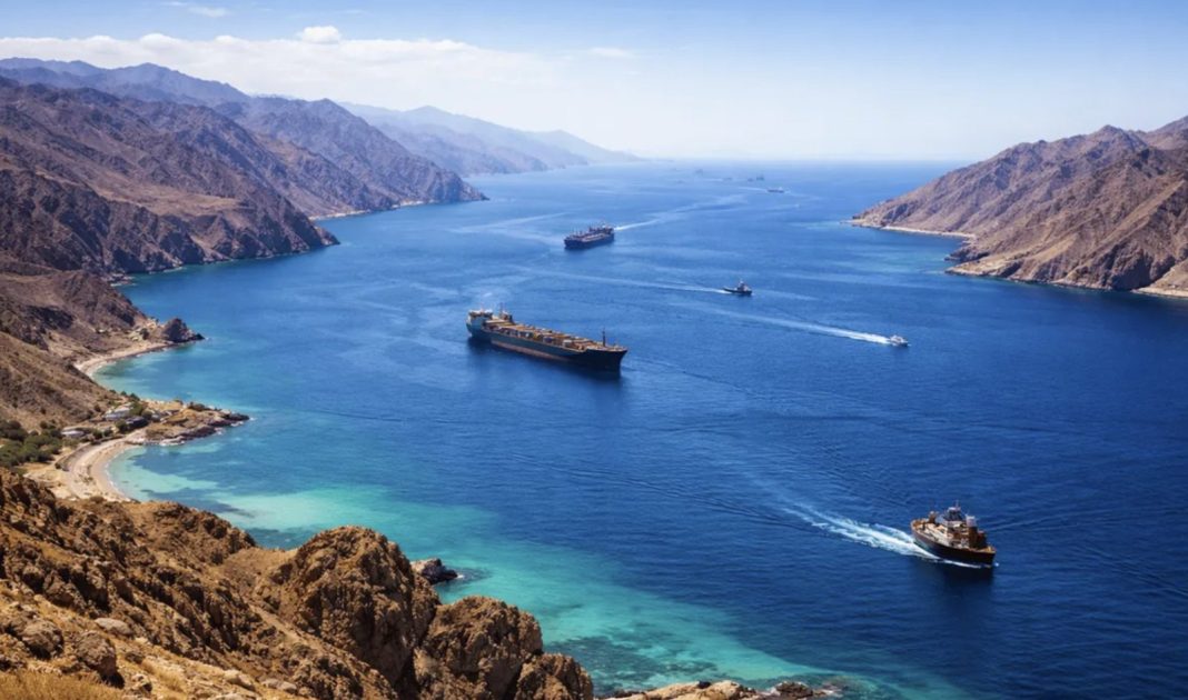 Calm blue sea with several cargo ships sailing between rugged brown mountains along a coastline.
