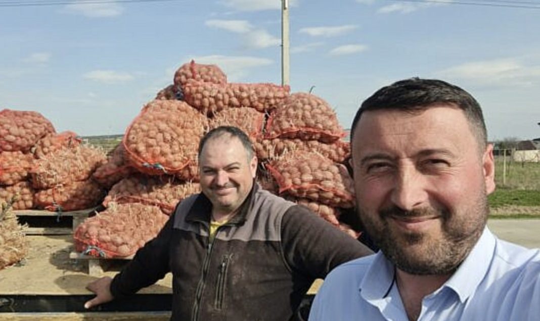 Two men pose and smile in front of large sacks of onions stacked outdoors on pallets in a rural setting.