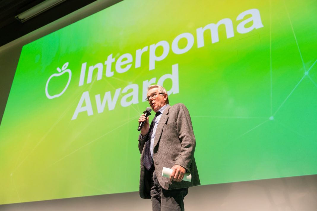 Man in a suit speaking into a microphone on stage, holding a rolled-up paper, with a bright green backdrop that reads 'Interpon a Award' behind him.
