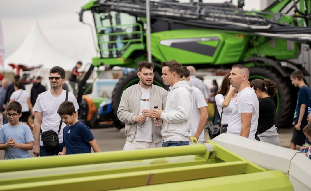 Group of people at an outdoor agricultural exhibition with a large green combine harvester behind them.