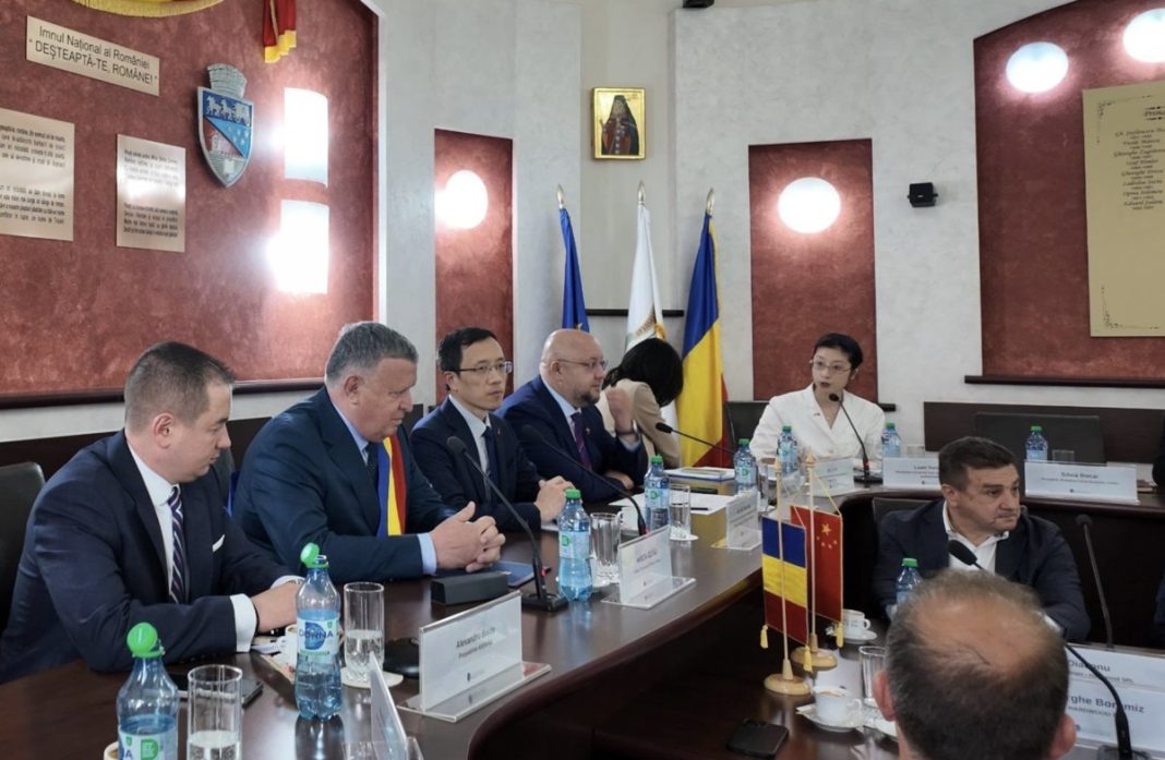 Panel of diplomats in a formal meeting room, seated at a long table with microphones, water bottles, and nameplates; flags stand behind them.