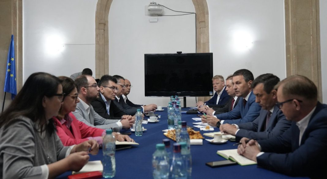 Business meeting with professionals in suits seated on both sides of a long conference table, water bottles and pastries in the center, European flag in the background.