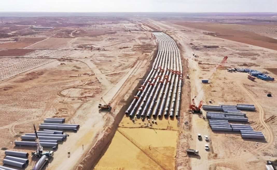 Aerial view of a desert pipeline project with many large steel pipes laid in trenches and construction equipment nearby.