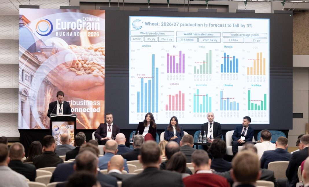 Conference stage with a speaker at a podium and a large screen displaying charts and world production data; audience seated in front.