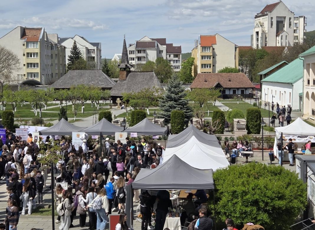 Outdoor market/fair in a town square with gray tents and a crowd of people, buildings visible in the background.