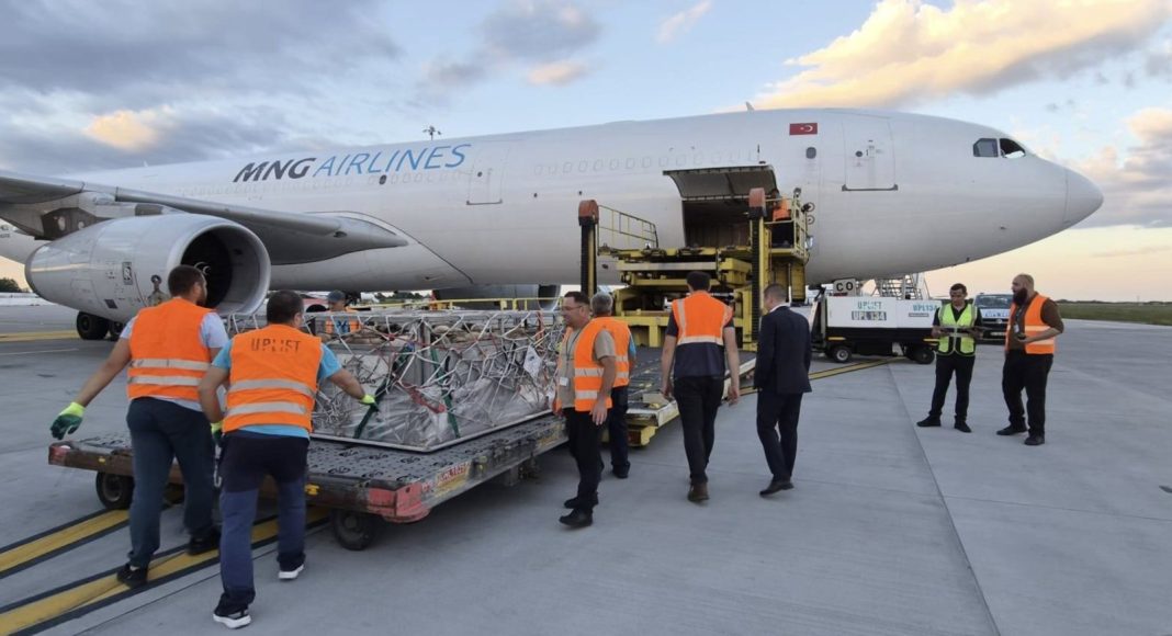 Ground crew in orange vests load a large cargo crate onto a white MNG Airlines plane on the tarmac with a forklift nearby.