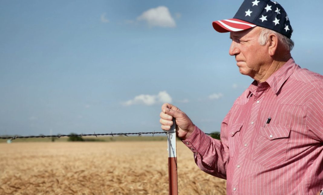 Older man in a red striped shirt and star-spangled cap stands beside a barbed-wire fence in a dry field.