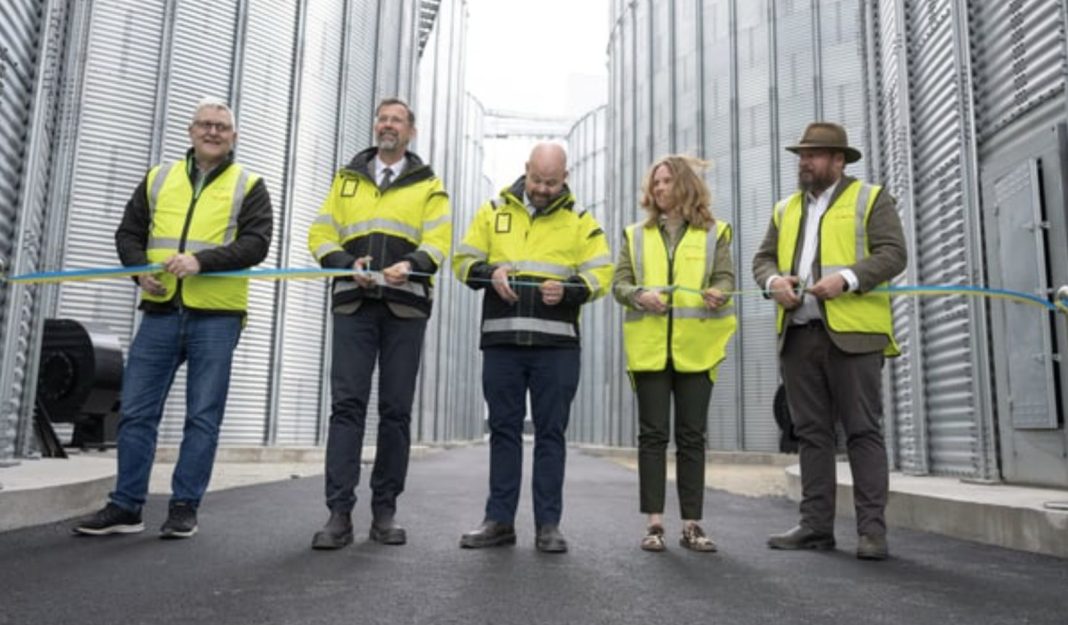 Five people in high-visibility vests cut a blue ribbon at an industrial site, marking a ribbon-cutting ceremony between silos.