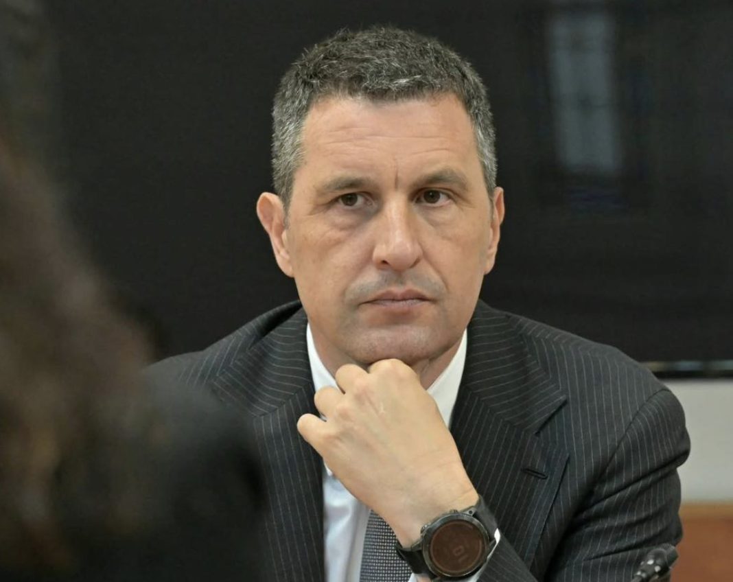 Man in a dark pinstriped suit rests his chin on his hand, looking serious in a meeting setting.”,