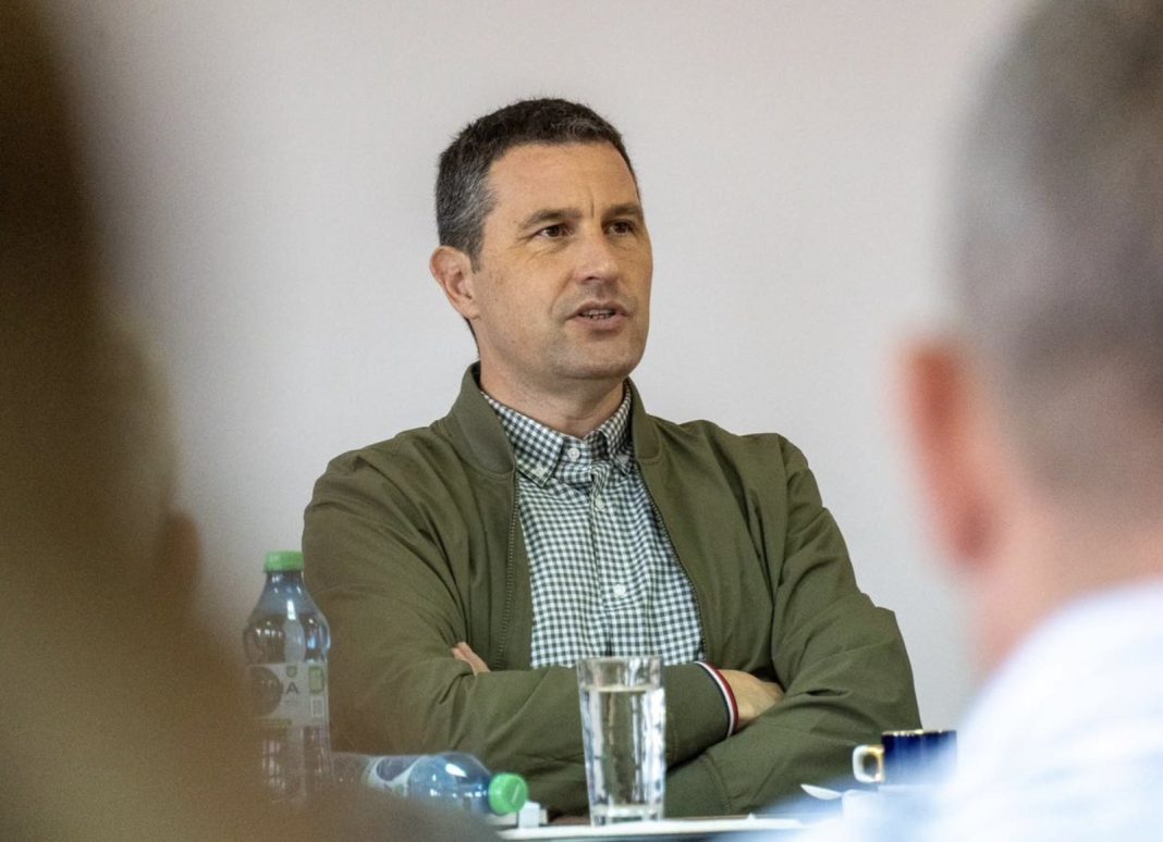 Man in a green jacket sits at a table during a meeting, speaking, with a water bottle and glass in front of him.