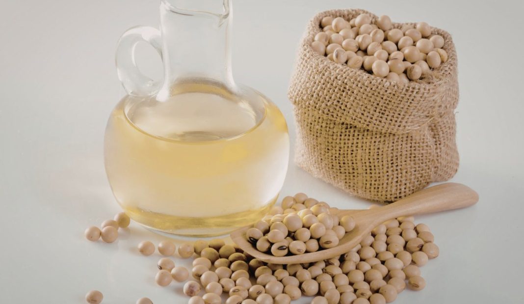Glass jug of light oil beside a burlap sack of soybeans and a wooden scoop, suggesting cooking oil from soybeans