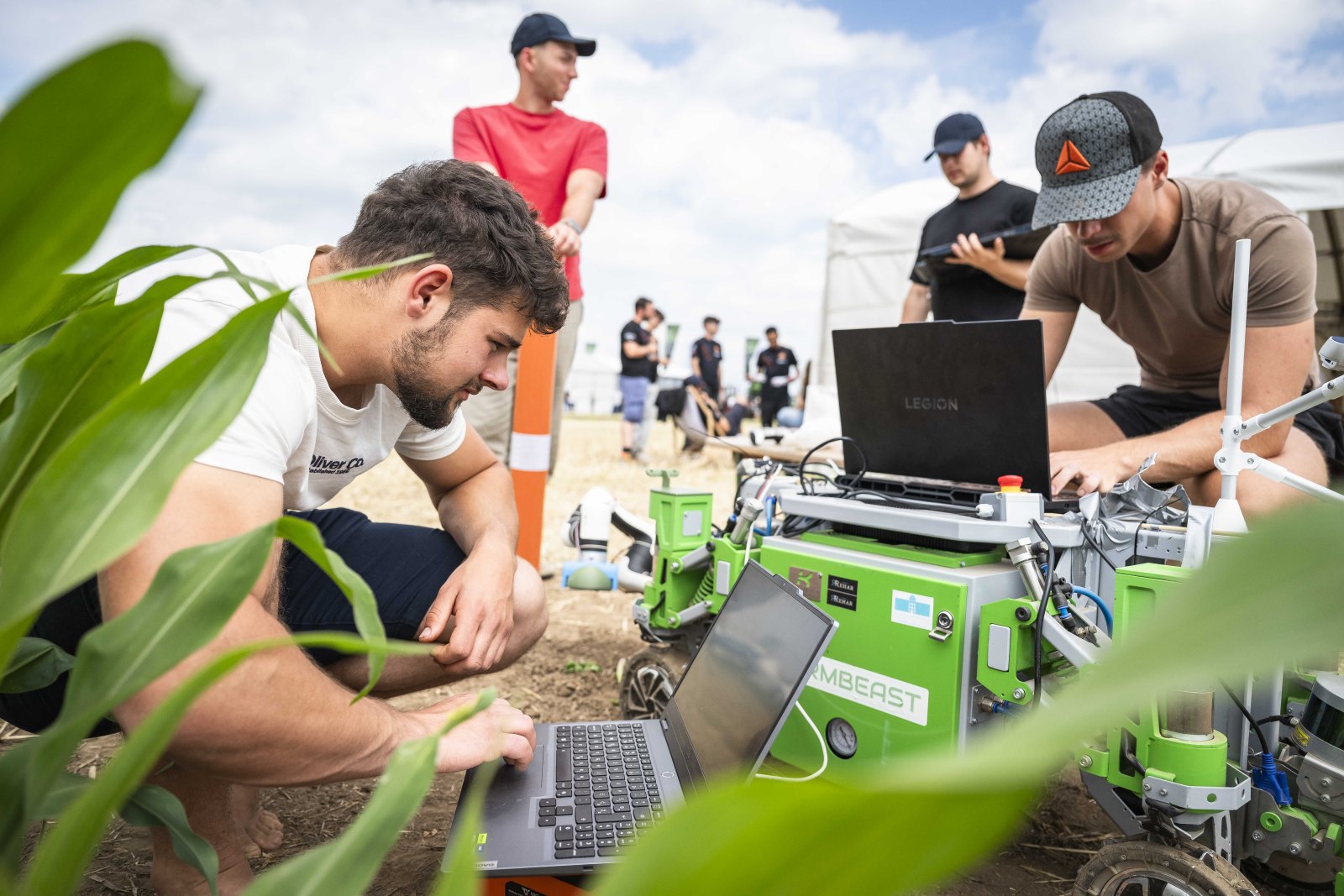 Two researchers test a green robotic device with laptops at an outdoor field lab, under a cloudy sky.