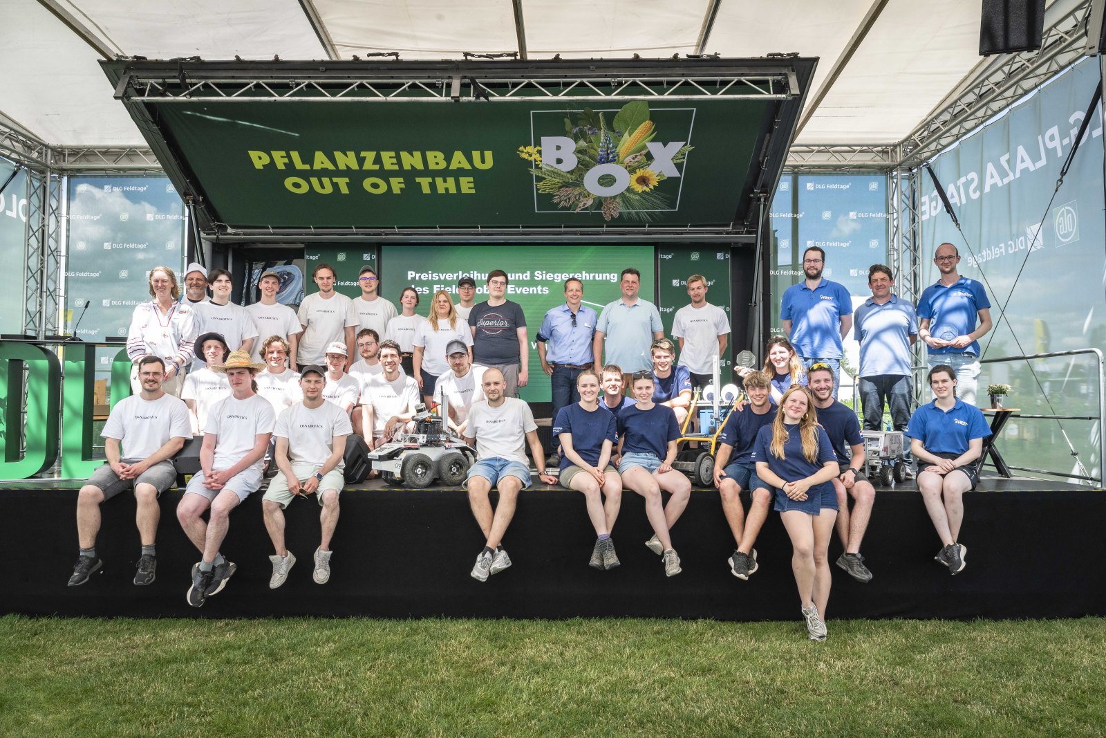 Group of about 40 people posing on an outdoor stage in front of a green banner that reads 'PFLANZENBAU OUT OF THE BOX', with stage equipment around them and a grassy foreground.