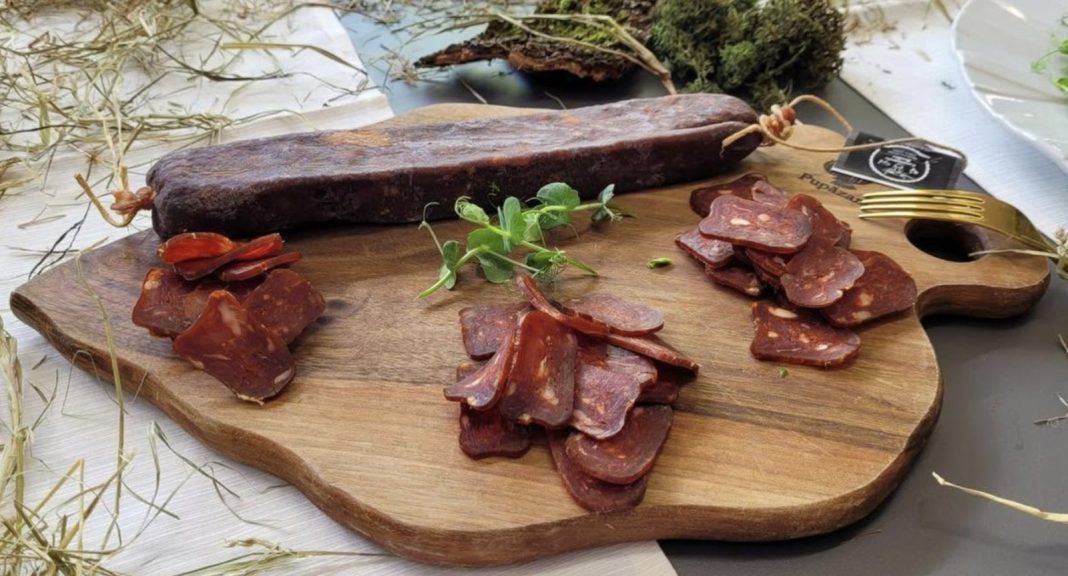 Cured meat slices on a wooden board with a whole smoked sausage, herbs, and a fork nearby in a rustic setting.