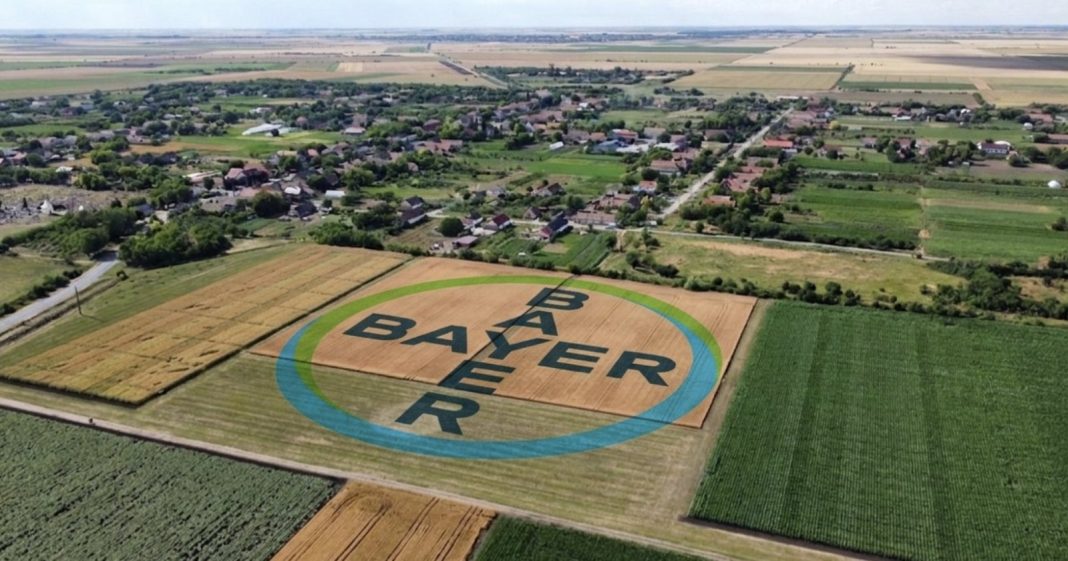 Aerial view of farmland with a large circular logo painted on a field reading 'BARBER' in bold letters, bordered by a blue-green ring near a village.
