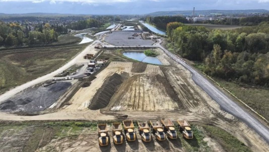 Aerial view of a large construction site with dirt piles, a row of dump trucks in the foreground, and winding roads through the area.