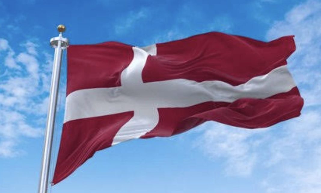 Danish flag (Dannebrog) waving on a flagpole against a blue sky.