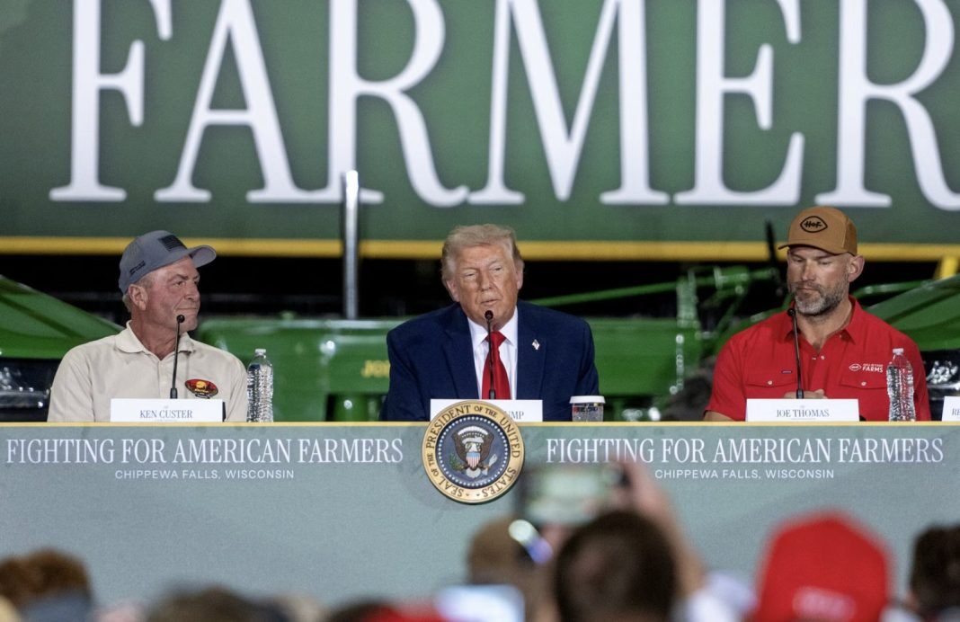 Panel at a farm event: a central speaker in a dark suit with red tie, flanked by two men, microphones in front, backdrop reading 'FARMER' and a banner 'Fighting for American Farmers, Chippewa Falls, Wisconsin'.