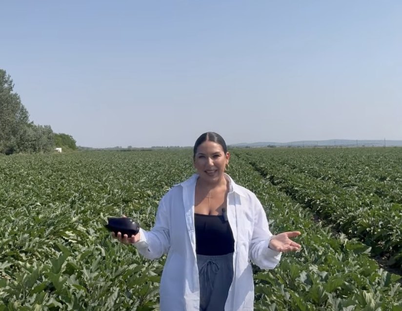 Woman in a white lab coat stands in a vast green field, holding an eggplant in one hand and gesturing with the other.