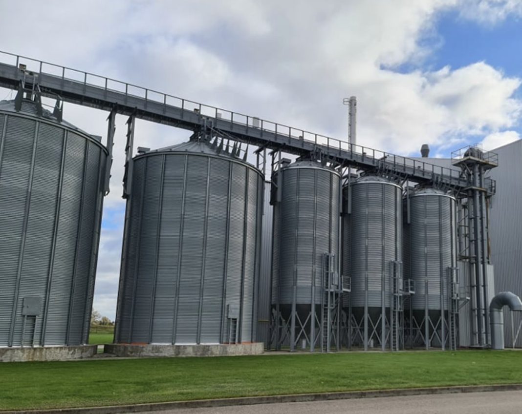 Row of tall galvanized grain silos with metal walkways and supports at an industrial facility, next to a grassy area under a partly cloudy sky