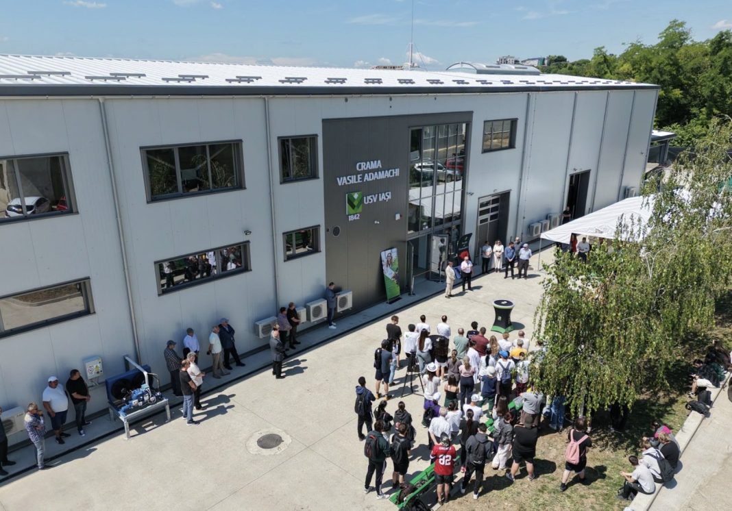 Aerial view of a gray industrial-style building with a crowd gathered outside for an outdoor ceremony, a speaker at a podium nearby and a white event tent on the right.