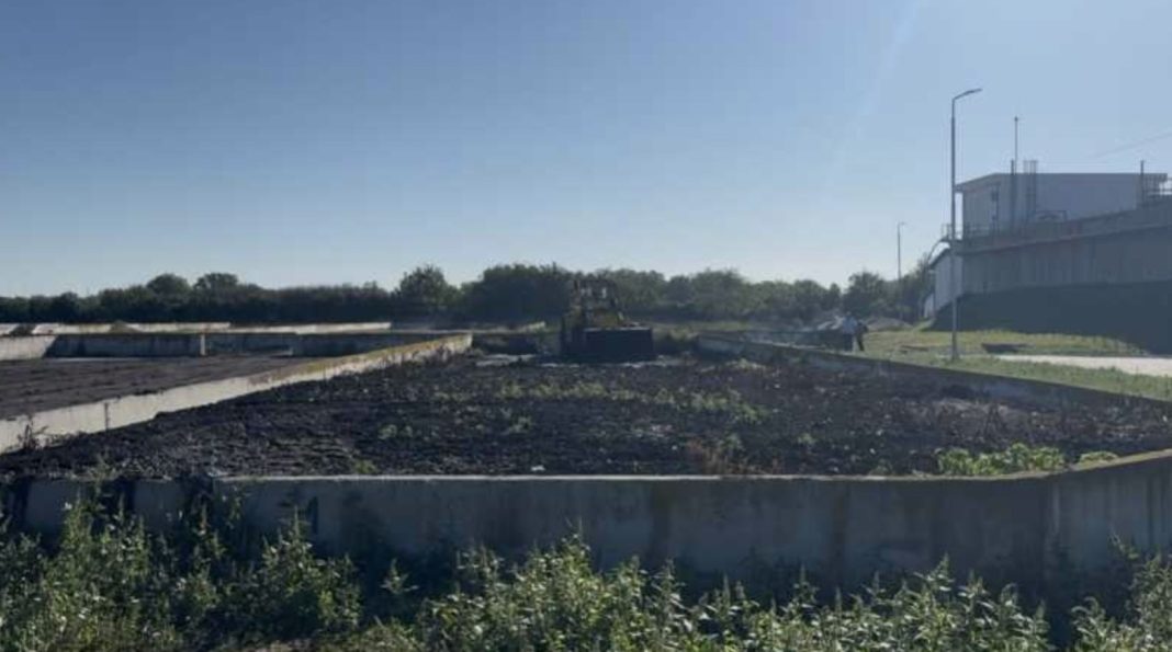 Construction site with a circular concrete trench, gravel and weeds, and a distant bulldozer near the treeline; a building and streetlights on the right.