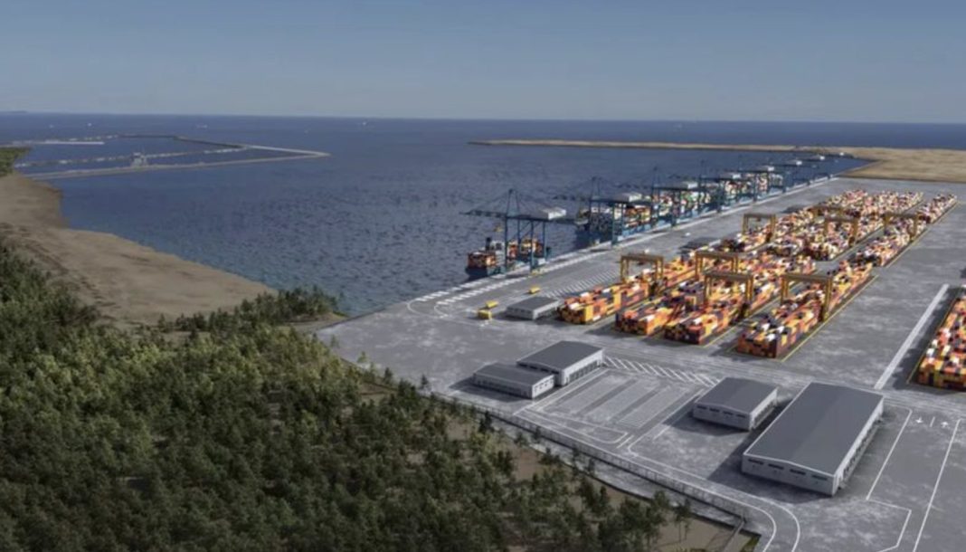 Industrial port with many stacked shipping containers, gantry cranes, and loading bays along the water, with a forested hillside in the foreground.