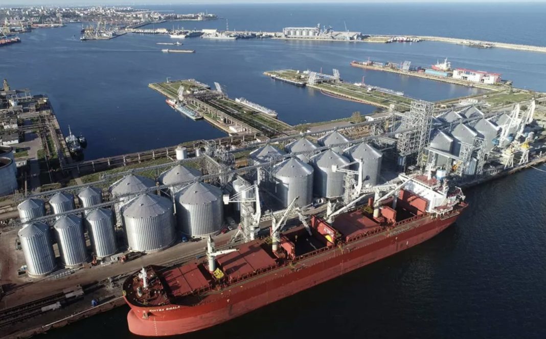 Aerial view of a busy port with a red cargo ship beside rows of silver silos and cranes at the dockyard