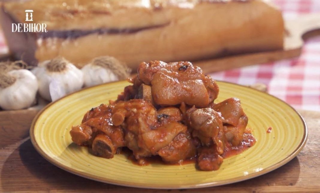 Plate of braised pork chunks in a tomato-based sauce on a yellow plate, with garlic and bread in the background and a De Bihor logo in the corner.