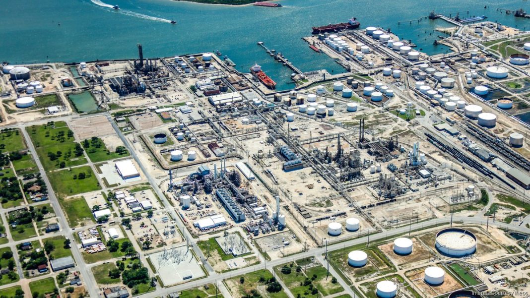A large oil refinery and storage tanks near a blue harbor, with networked pipes, cranes, and adjacent roads and green areas.