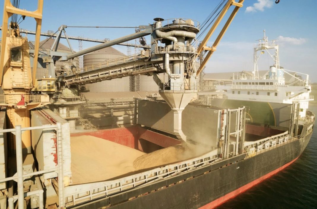 Bulk carrier being loaded/unloaded with a conveyor system at an industrial grain/sand terminal; silos and cranes in the background reveal a large port facility.