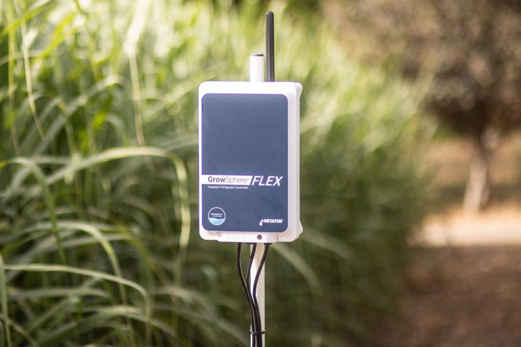 GrowSphere FLEX irrigation controller mounted on a stake in a garden with green plants behind it.