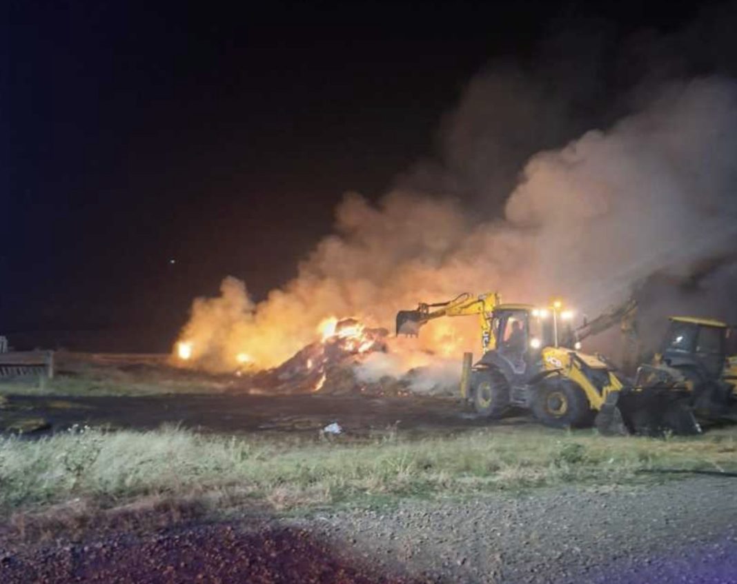 Night scene of a large fire burning a pile of debris next to yellow construction equipment; thick smoke rises at night.