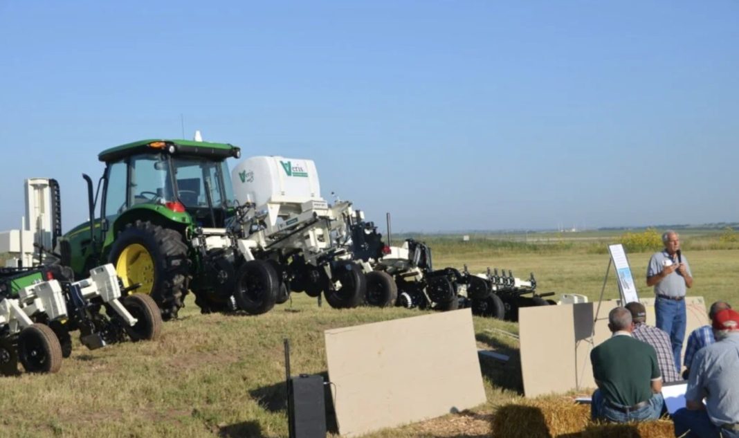Green tractor connected to large farming equipment on a sunny field, with a speaker addressing a small audience nearby.