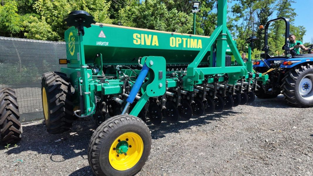 Green SIVA OPTIMA seed drill attached to a blue tractor on a gravel lot, outdoors