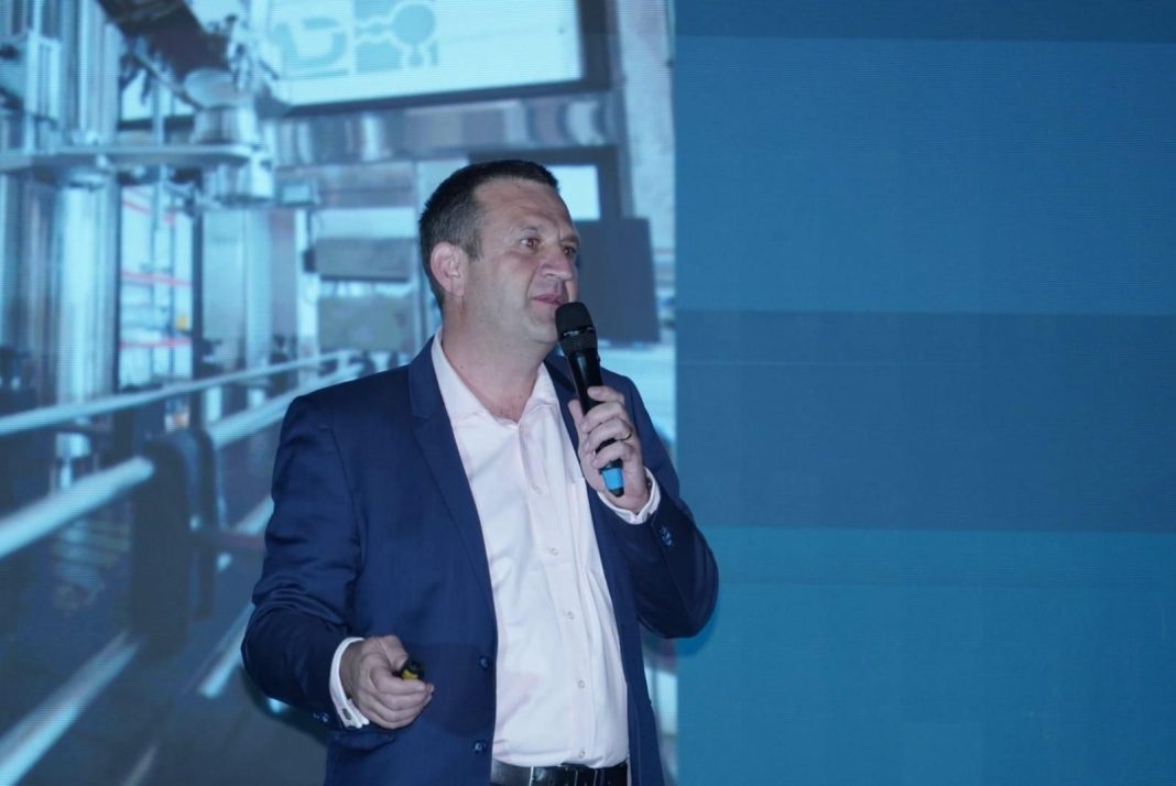 Man in a navy suit speaking into a handheld microphone on stage, with a blue screen backdrop behind him.